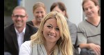 Portrait einer blonden Frau, im Hintergrund sind schemenhaft vier andere Personen zu erkennen